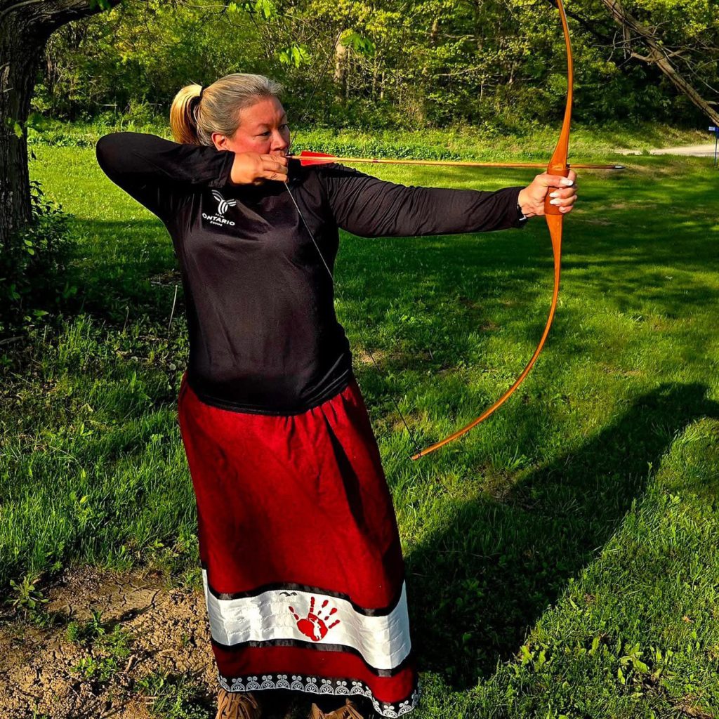 An indigenous woman in a red ribbon dress with her bow and arrow displaying her archery expertise.